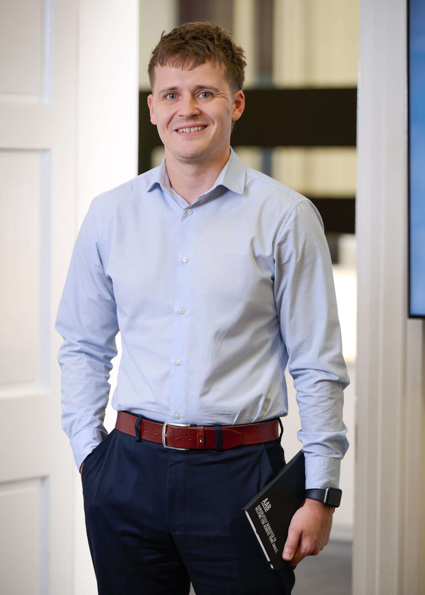 An image of James Taylor wearing a blue shirt with black trousers. He is standing in the office holding a notebook.