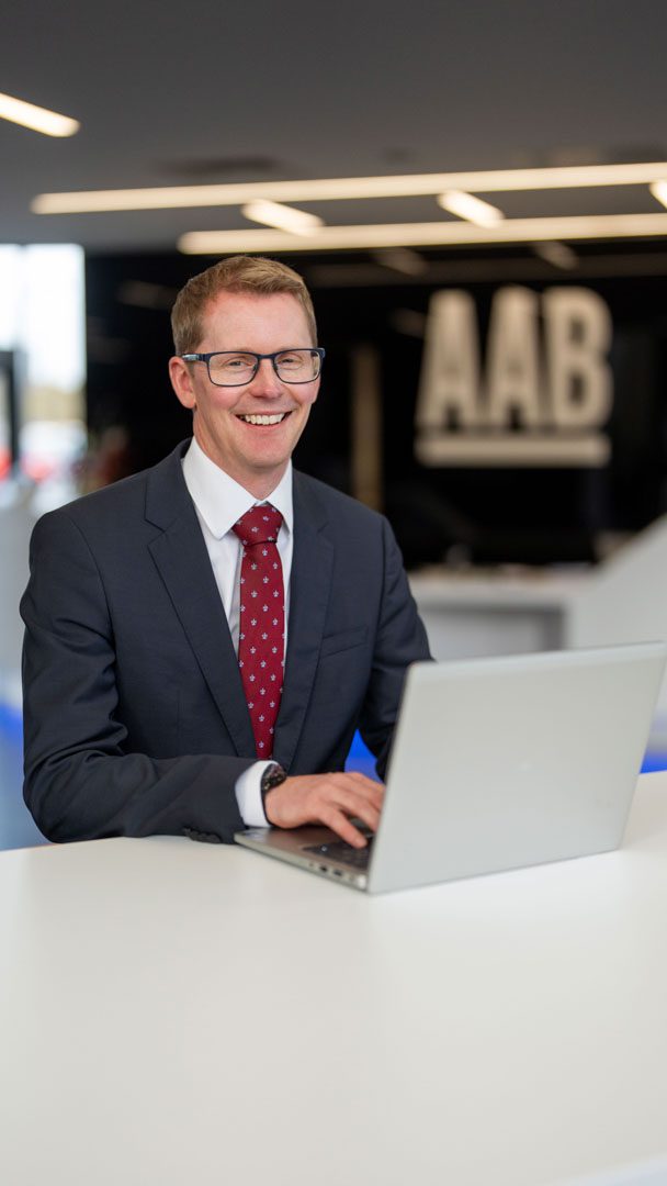 An image of Andrew Webster sitting at an office desk wearing a grey suit jacket, burgundy tie. He is working on his laptop and smiling at the camera.