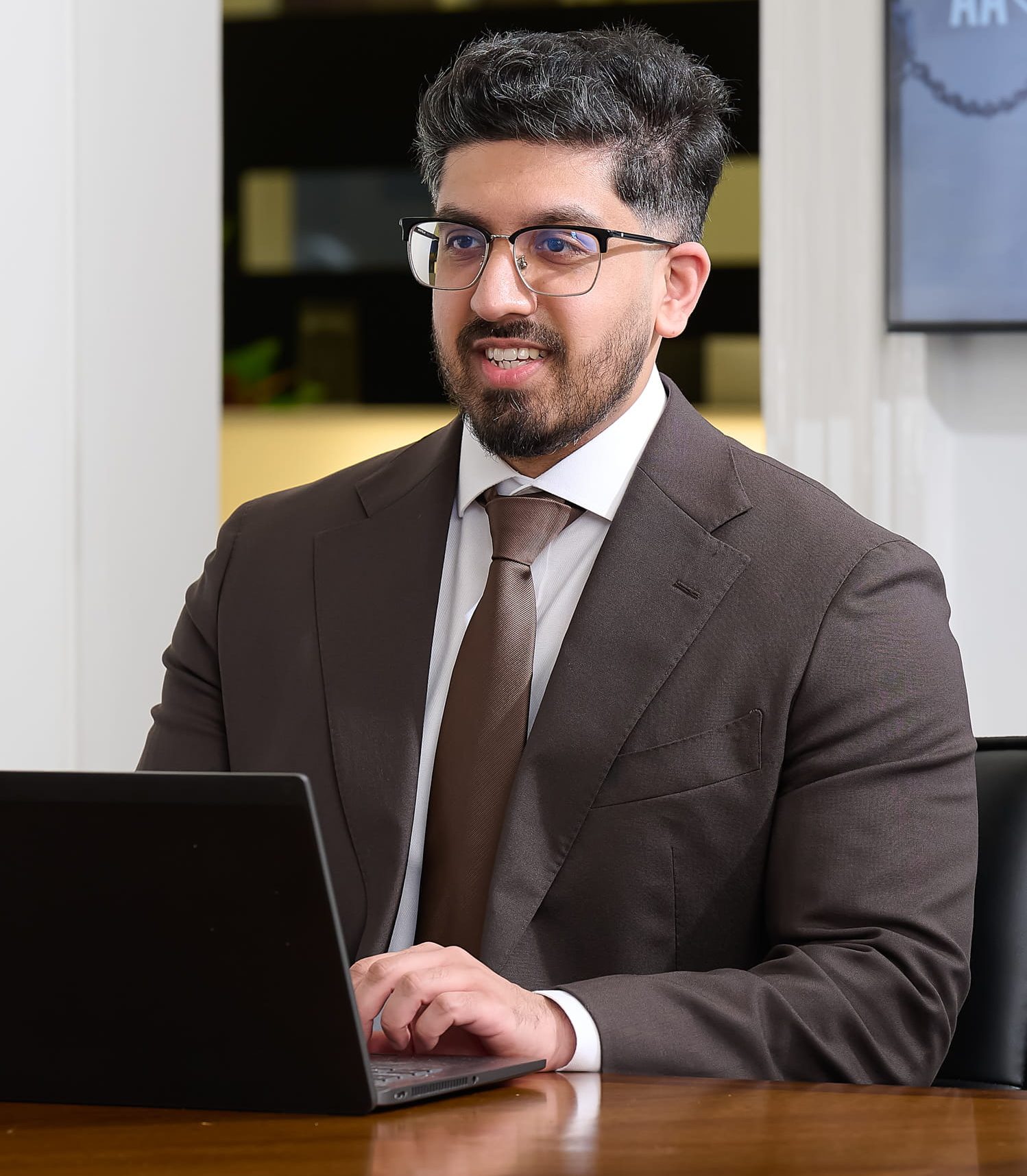 An image of Rafiq Gazi wearing a brown jacket and brown tie. He is sitting at a desk with his laptop.