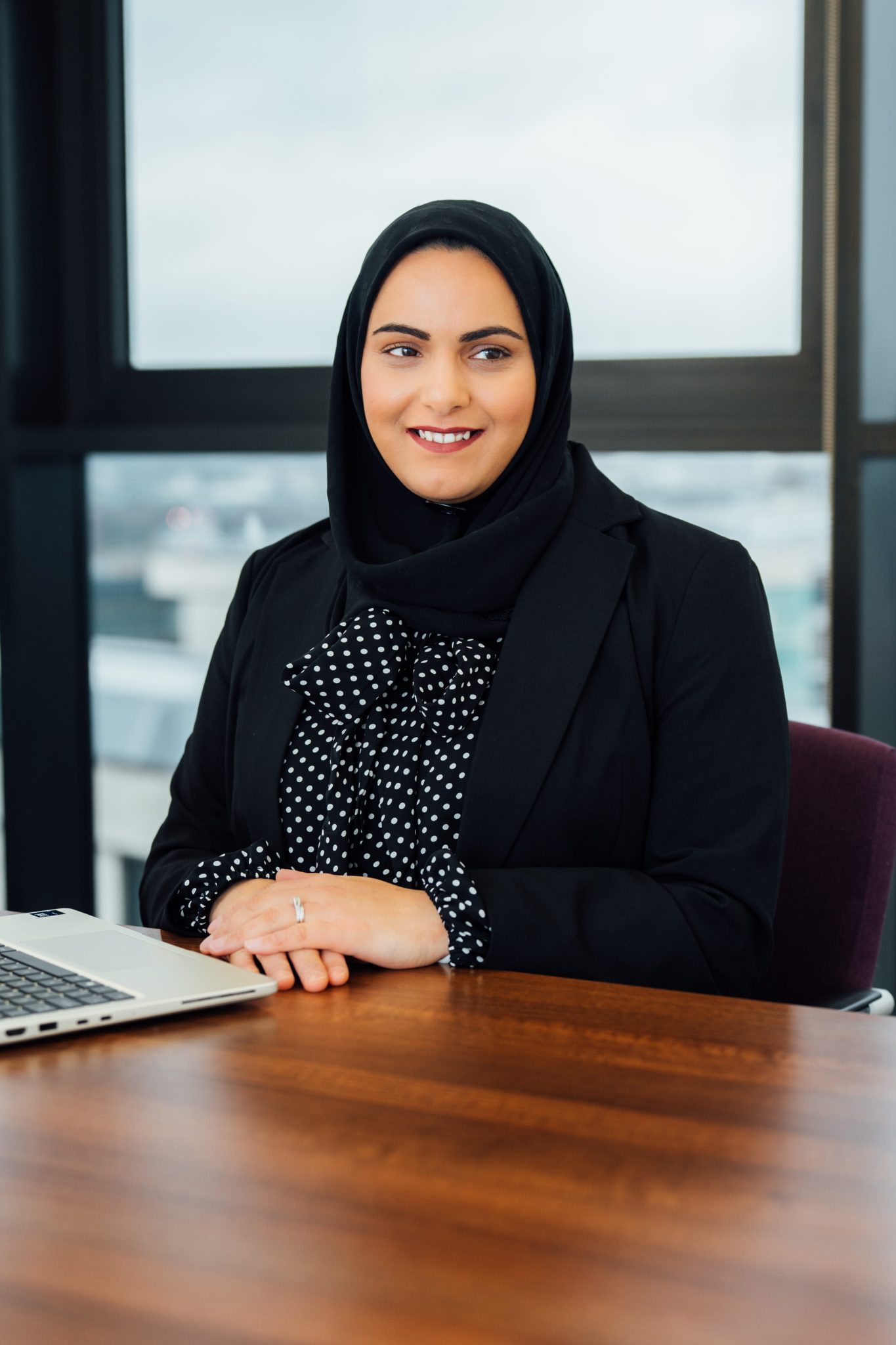An image of Aishah Akhtar sitting at a desk, on her laptop and smiling at the camera.